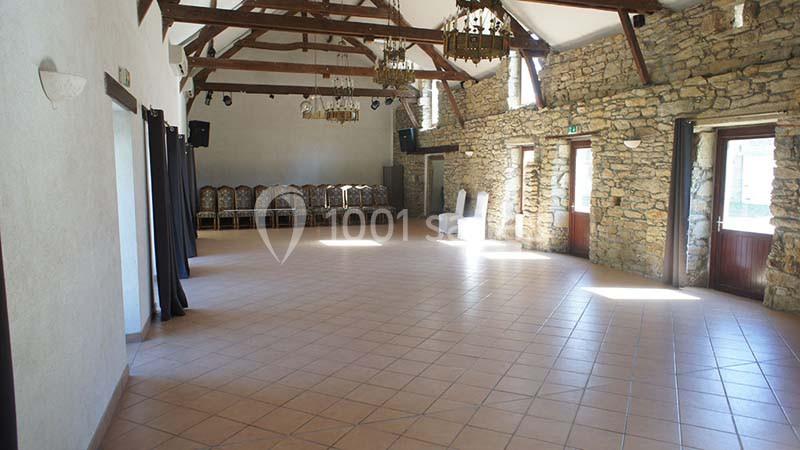 Salle spacieuse avec murs en pierre, poutres apparentes, sol carrelé et chaises empilées au fond.