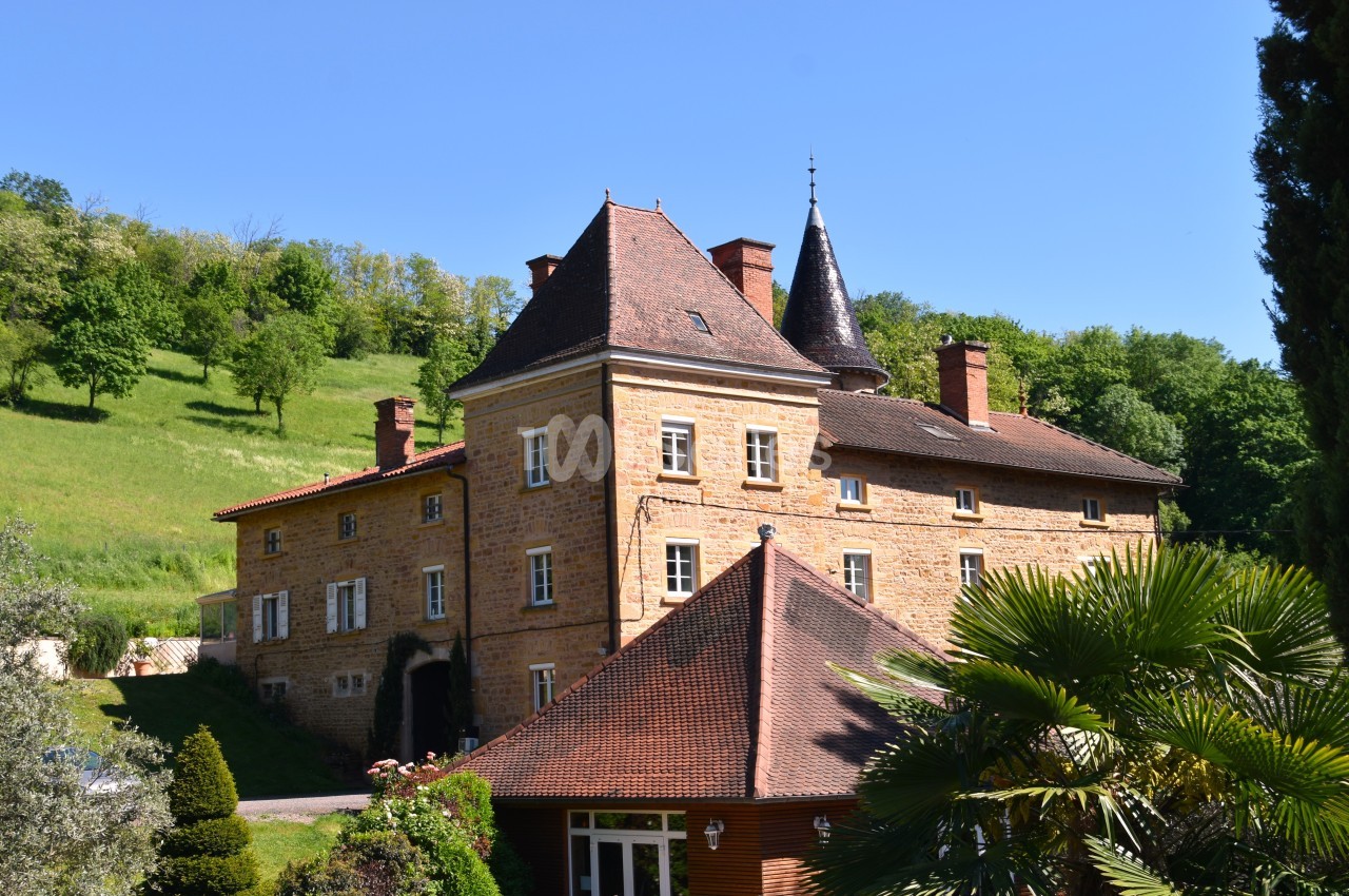 Grande maison en pierre avec tour, entourée de verdure et située dans un paysage rural ensoleillé.