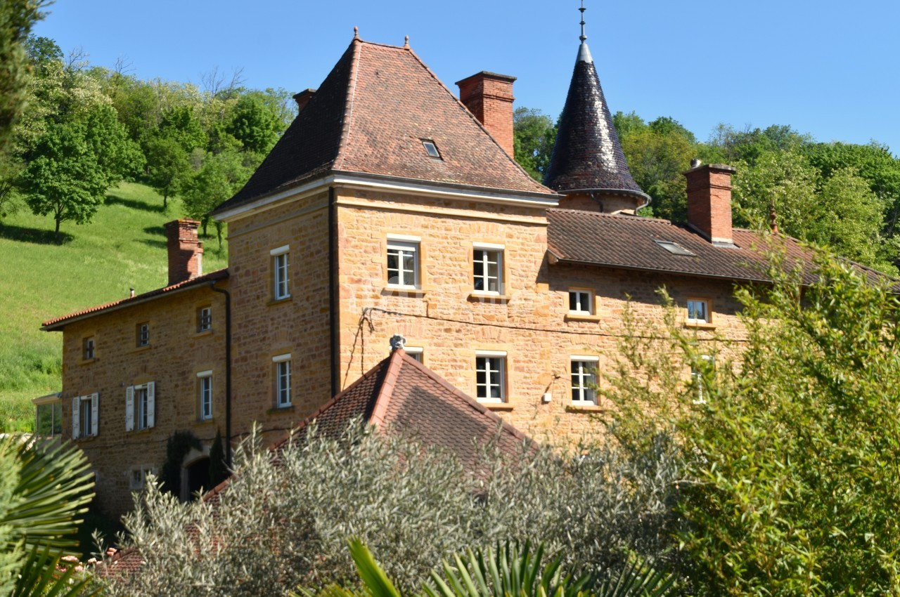Grande maison en pierre avec toit en tuiles, tour conique et végétation environnante sous un ciel bleu.
