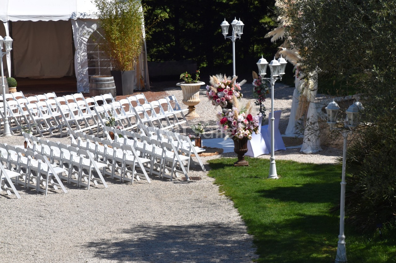 Chaises blanches alignées en extérieur pour une cérémonie, avec décorations florales et tente en arrière-plan.