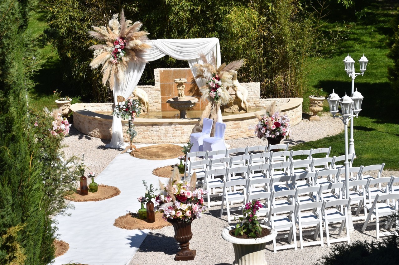 Allée centrale décorée menant à une arche de mariage ornée de fleurs et de voilages, entourée de chaises blanches.