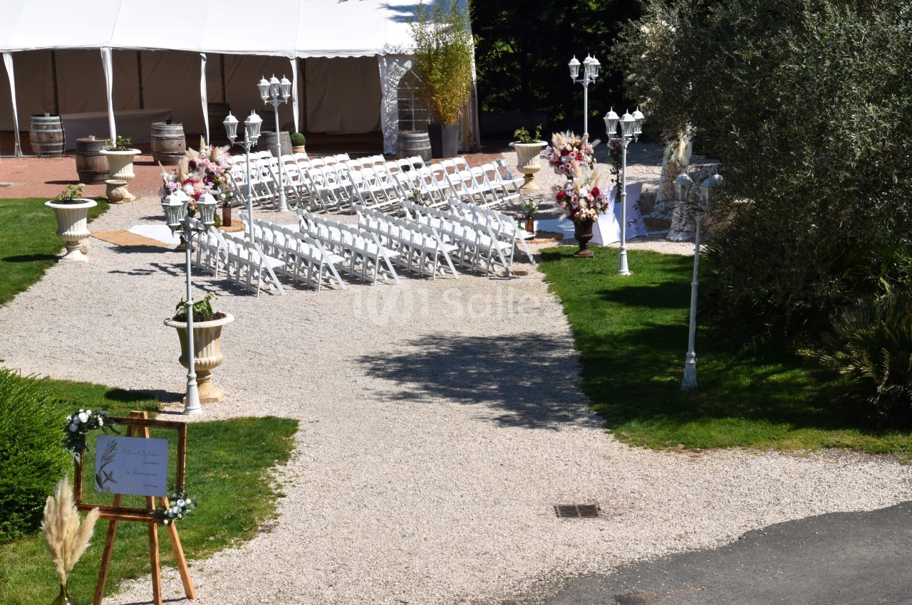 Allée gravillonnée menant à une cérémonie en plein air avec chaises blanches, décorations florales et tente blanche.