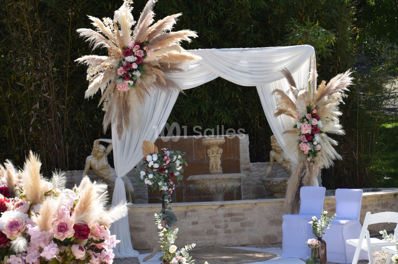 Arche de cérémonie décorée de voilages blancs, fleurs roses et pampas, devant une fontaine en pierre.