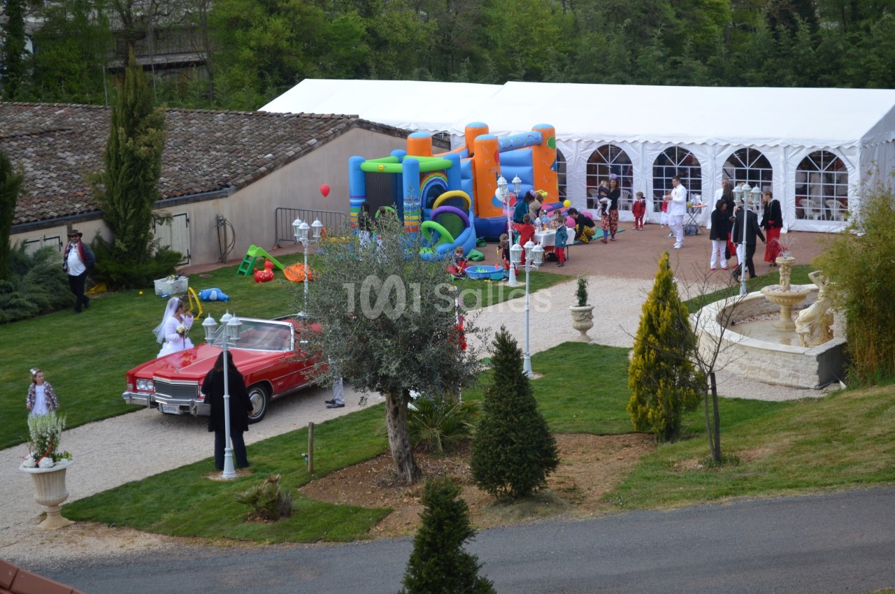 Vue d'une fête en extérieur avec une voiture rouge, château gonflable, invités et une tente blanche.