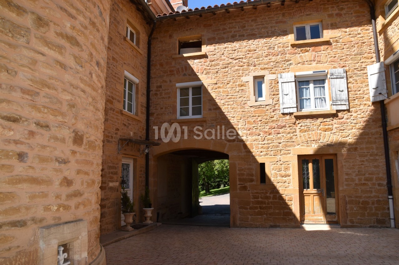 Cour intérieure en pierre dorée avec une arche menant à un espace ouvert, entourée de fenêtres et volets en bois.