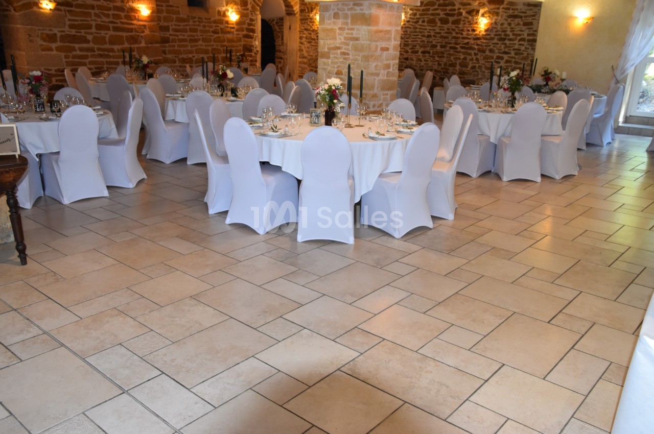 Salle de réception décorée avec des tables rondes dressées, nappes blanches et chaises houssées, éclairage chaleureux.