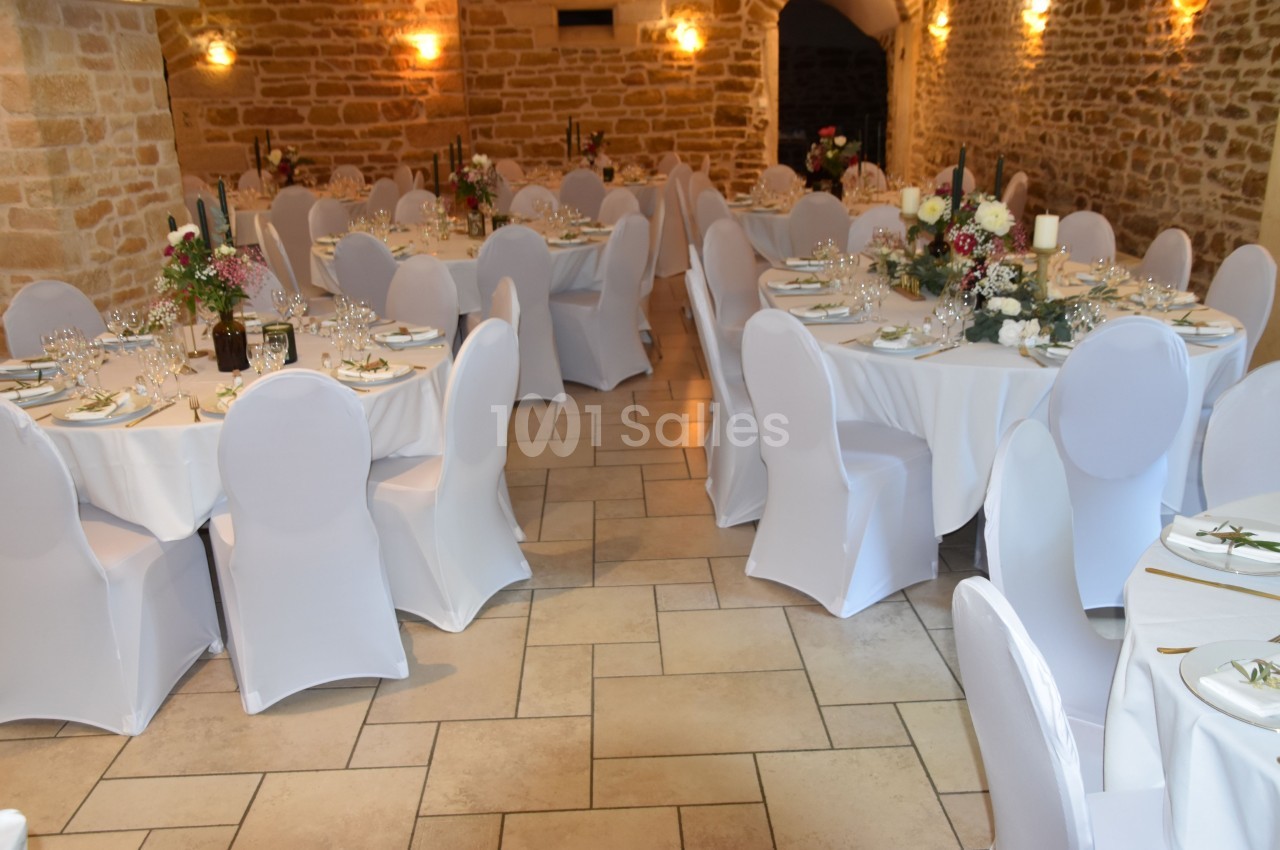 Salle de réception en pierre avec tables rondes dressées, nappes blanches et décorations florales.