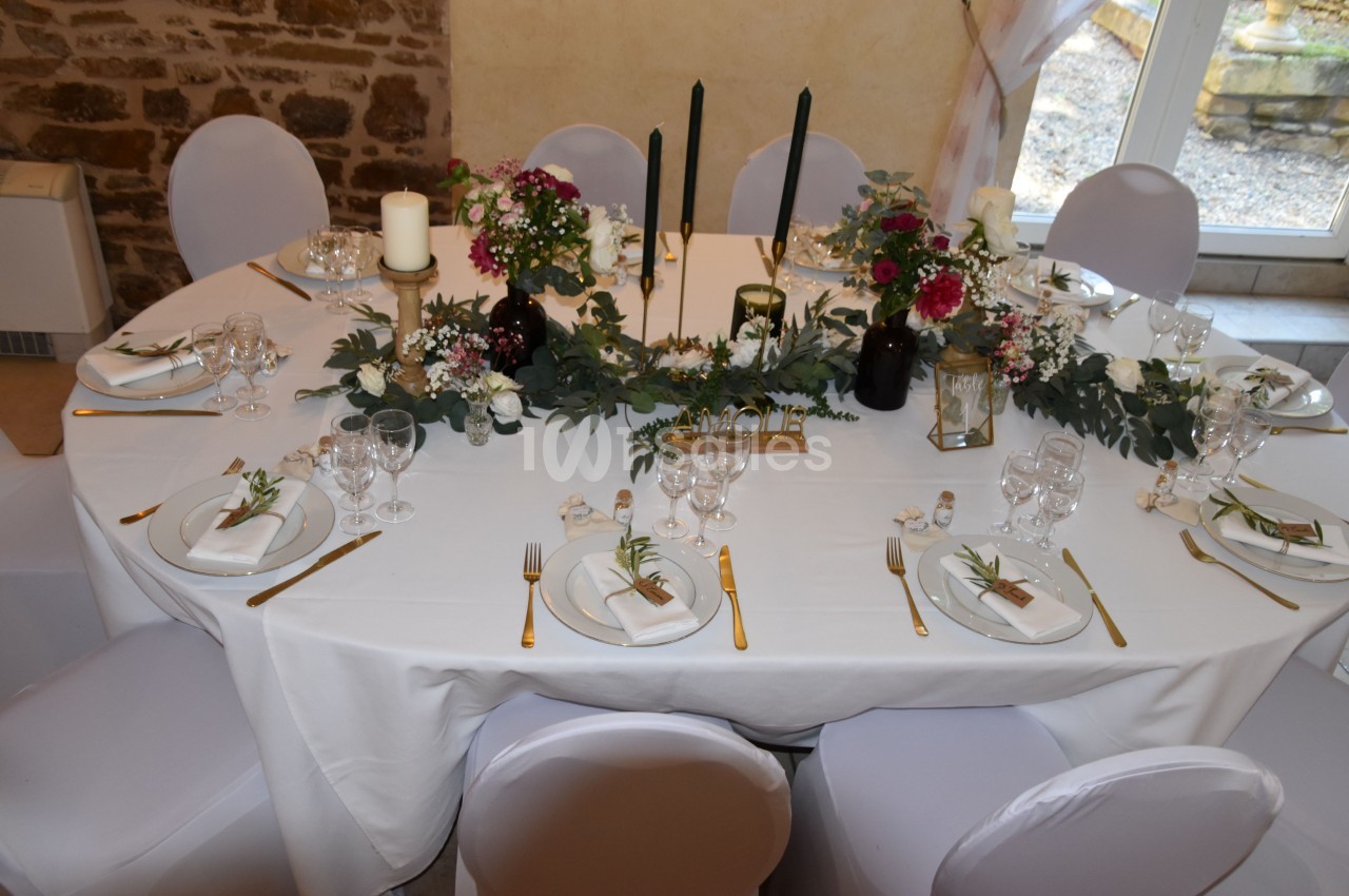 Table ronde élégamment dressée avec nappes blanches, bougies, fleurs et couverts dorés dans une salle en pierre.