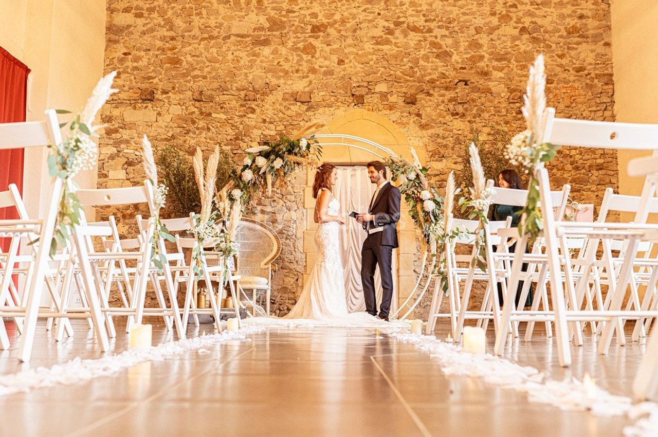 Un couple échange ses vœux dans une salle en pierre décorée d'arceaux fleuris et de chaises blanches.