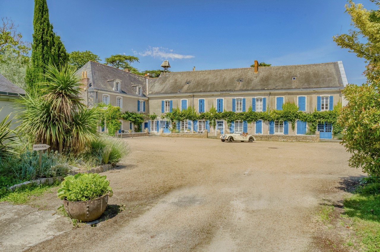 Cour d'une grande maison en pierre avec volets bleus, entourée de végétation et d'un ciel dégagé.