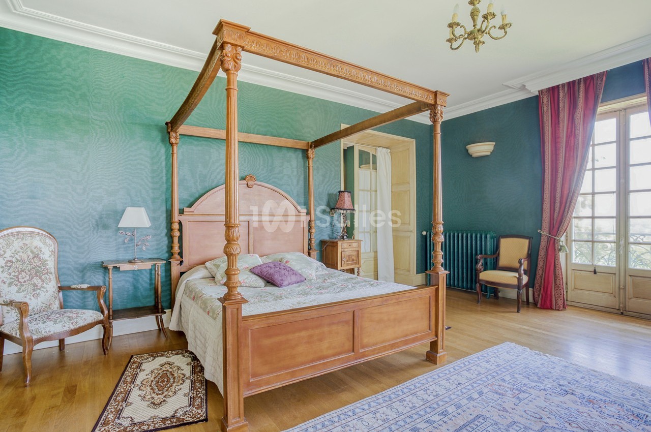 Chambre avec lit à baldaquin en bois, murs verts, parquet, tapis, fauteuils et grande fenêtre avec rideaux rouges.