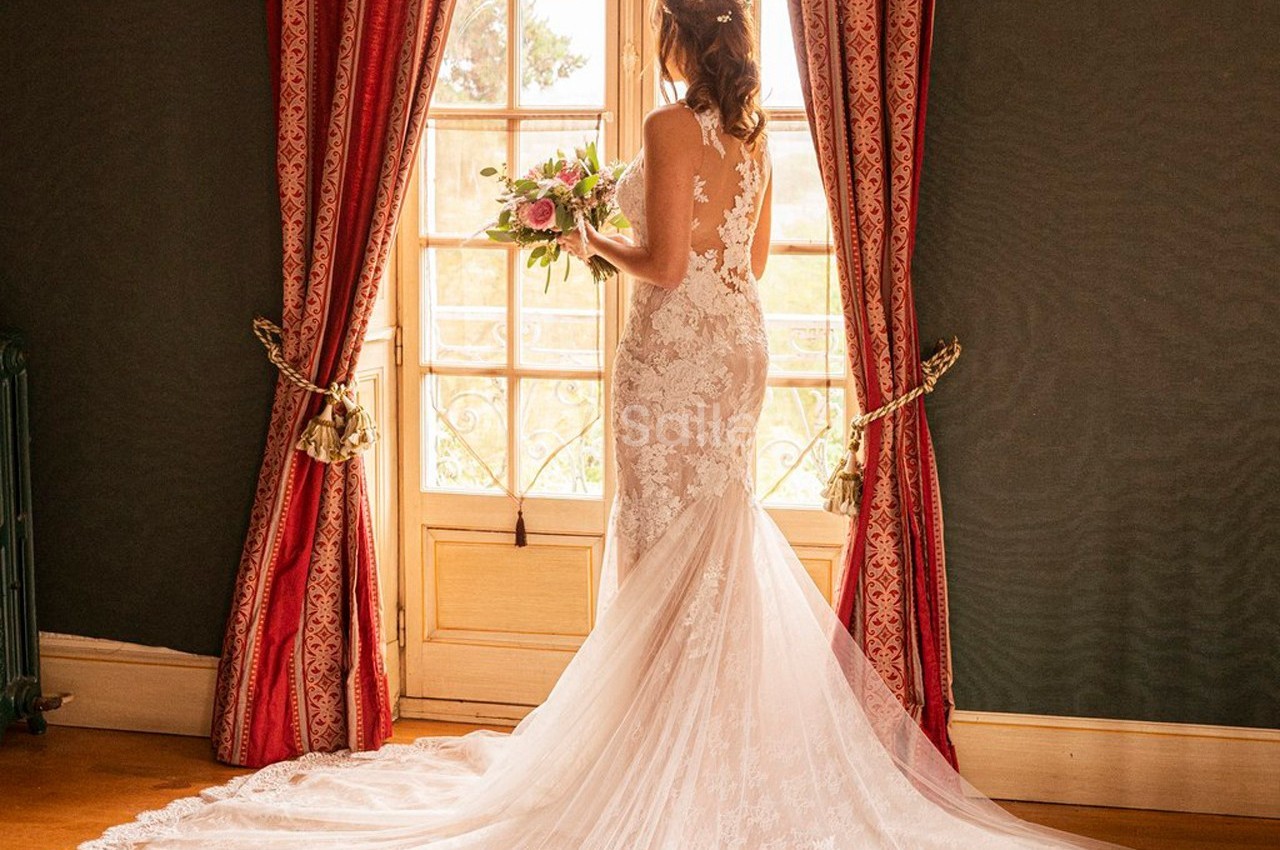 Une femme en robe de mariée en dentelle regarde par une fenêtre encadrée de rideaux rouges dans une pièce élégante.