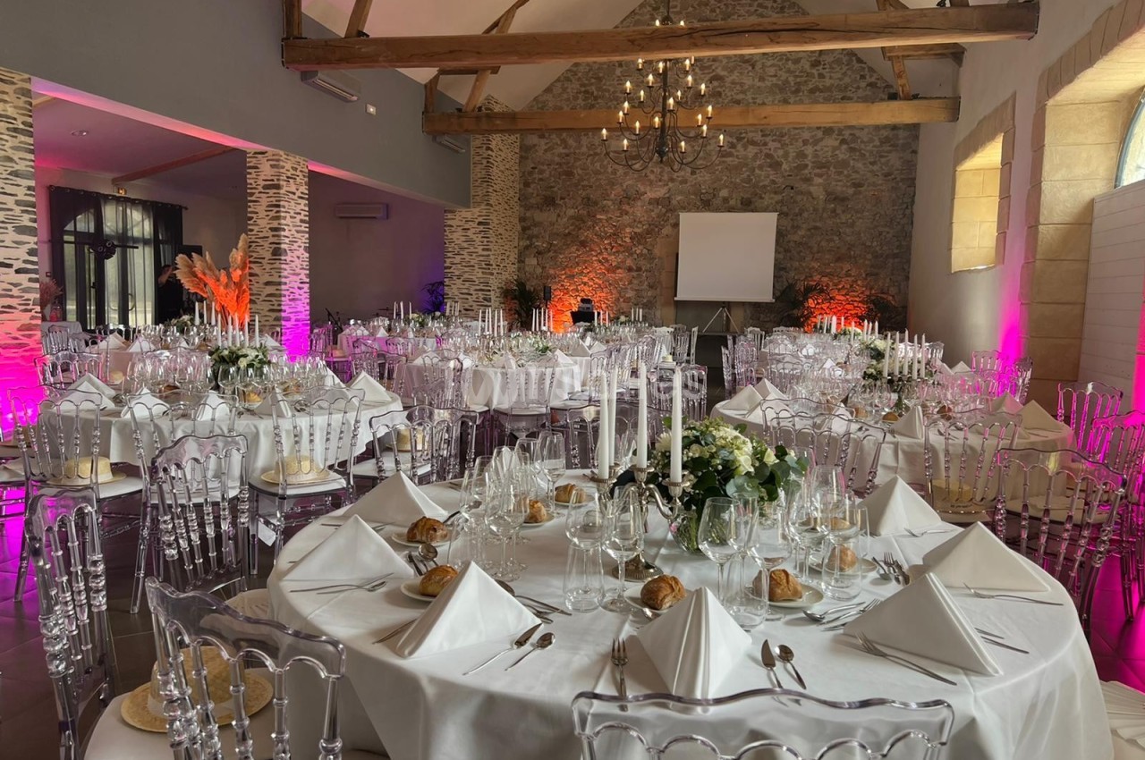 Salle de réception décorée avec des tables rondes élégantes, nappes blanches et chaises transparentes sous des poutres…