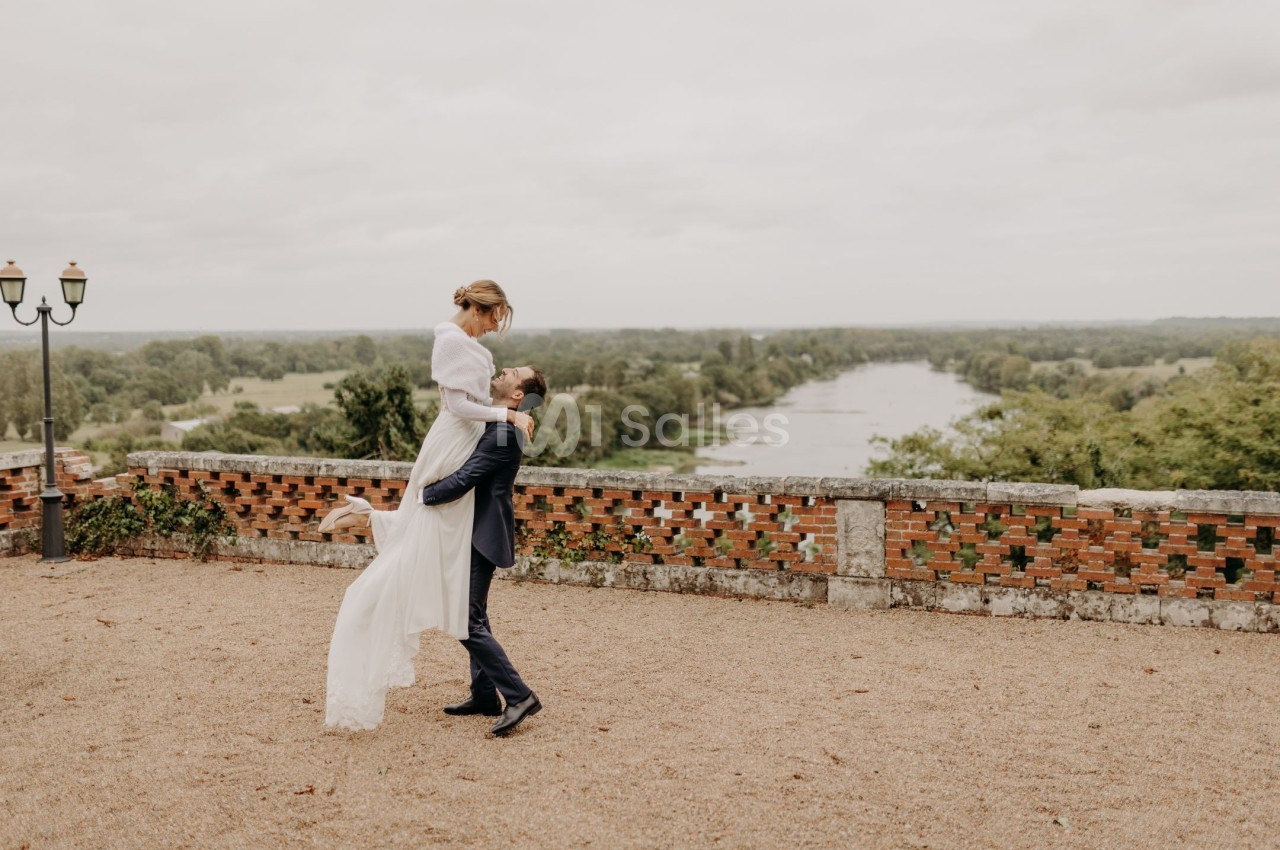 Un homme en costume soulève une femme en robe blanche dans une cour avec vue sur une rivière et des arbres.
