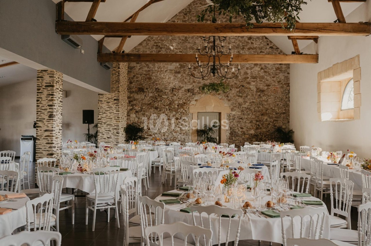 Salle de réception lumineuse avec des tables rondes dressées, des chaises blanches et des murs en pierre apparente.