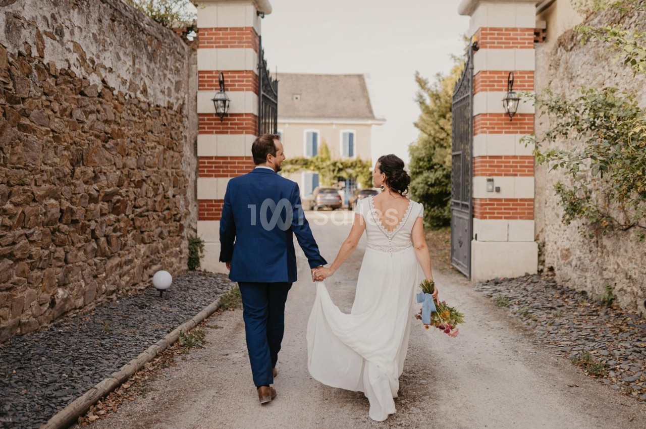 Un couple en tenue de mariage marche main dans la main sur une allée bordée de murs en pierre et en briques.