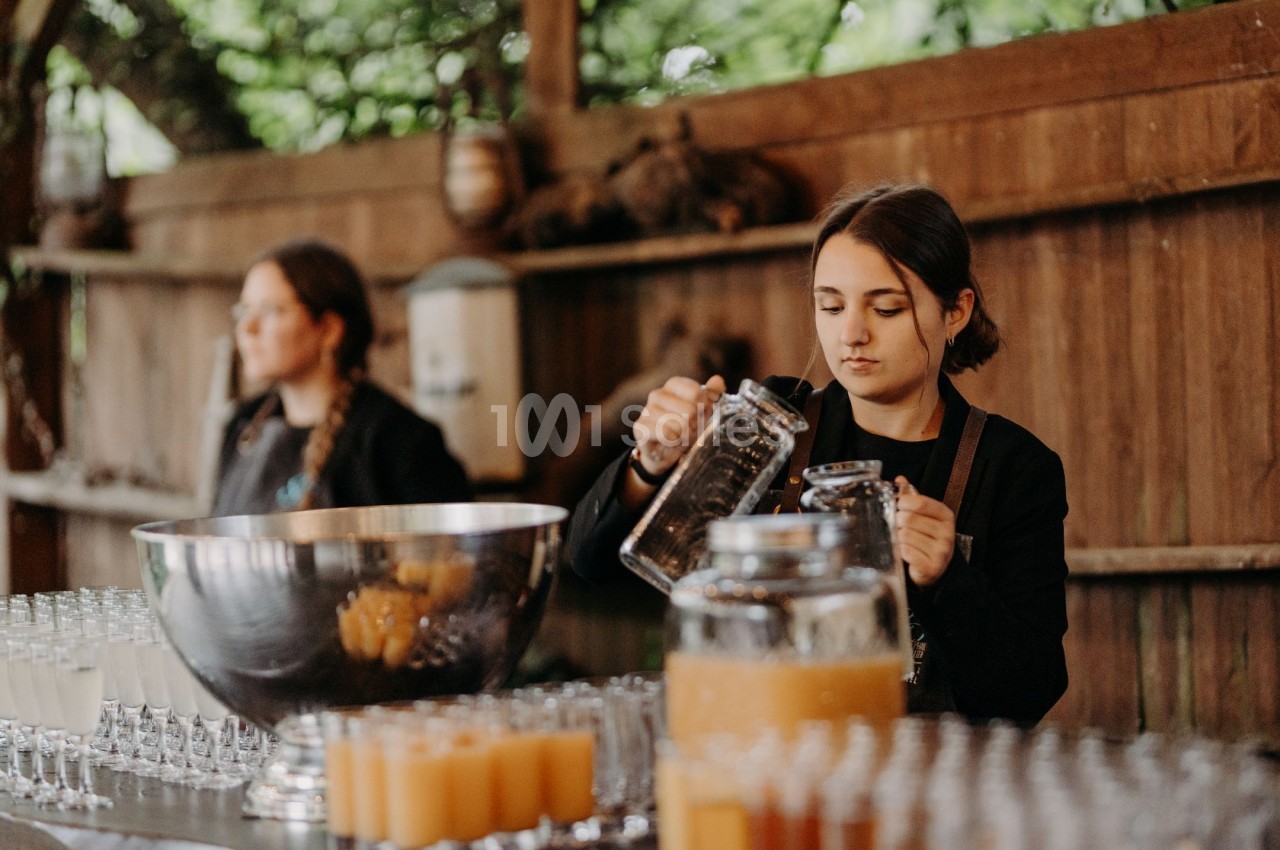 Une femme verse une boisson dans un verre sur un comptoir rempli de verres et de carafes, en extérieur.