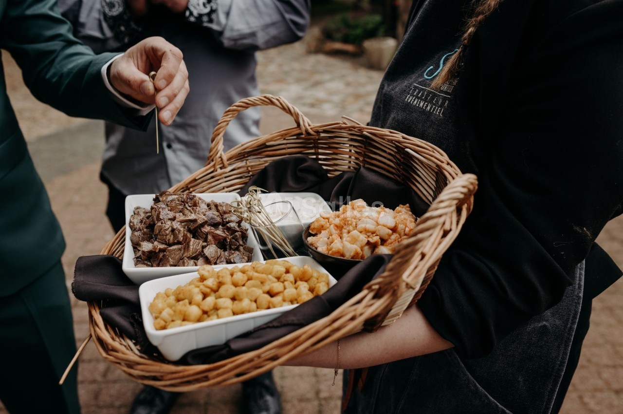 Plateau en osier contenant des bols de viande, crevettes et croquettes, tenu par une personne en uniforme.