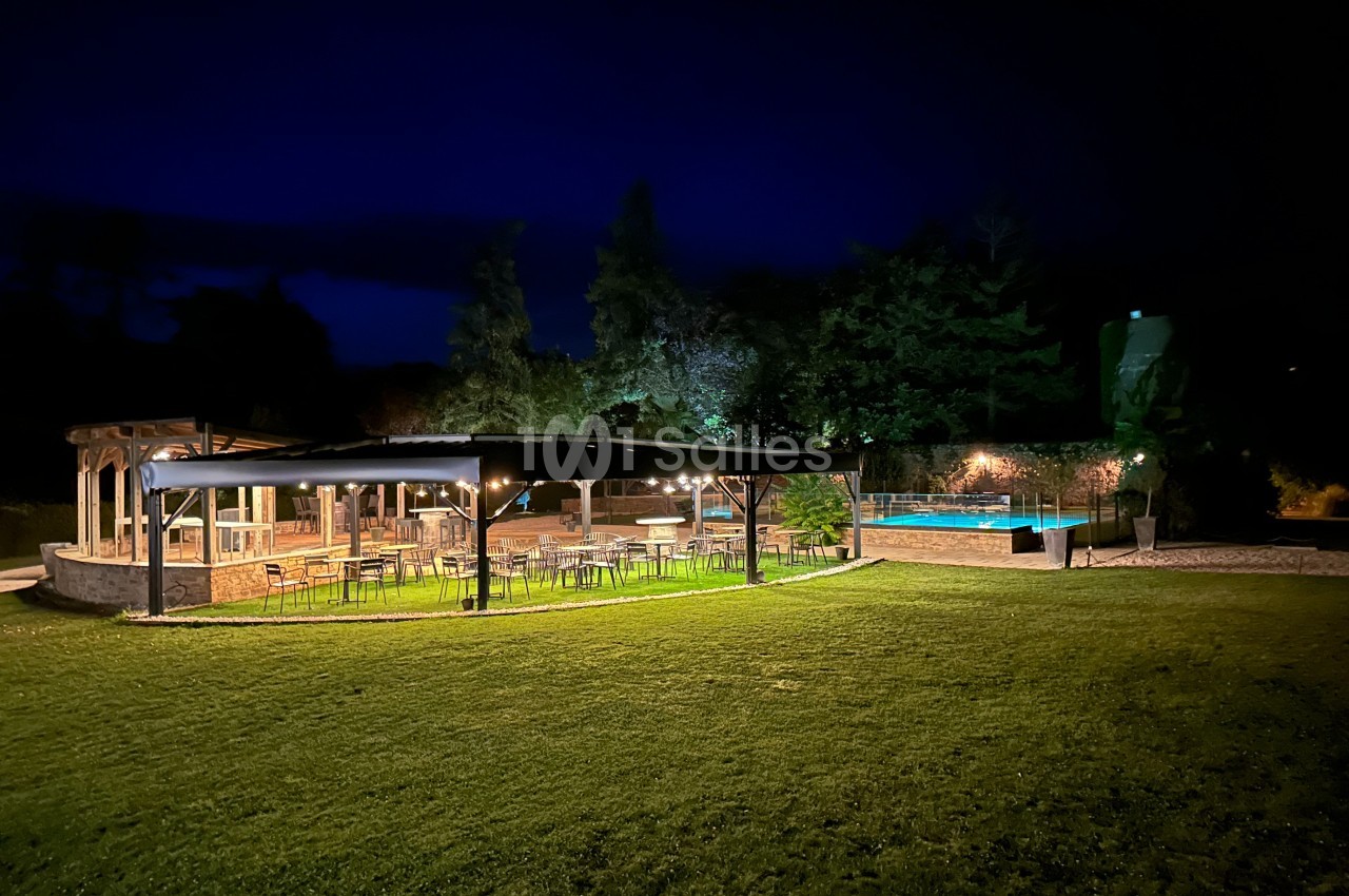 Espace extérieur éclairé la nuit avec terrasse couverte, tables, chaises et piscine entourée de verdure.