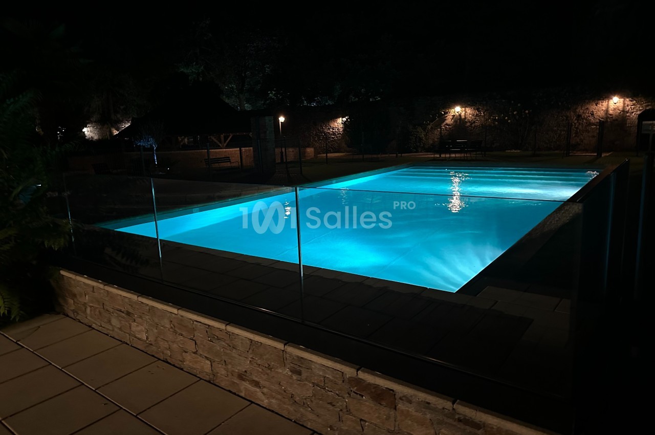 Piscine éclairée de nuit entourée de végétation et d'un muret en pierre, avec un éclairage tamisé en arrière-plan.