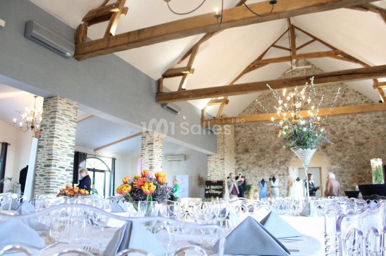 Salle de réception lumineuse avec poutres apparentes, tables dressées et décoration florale élégante.