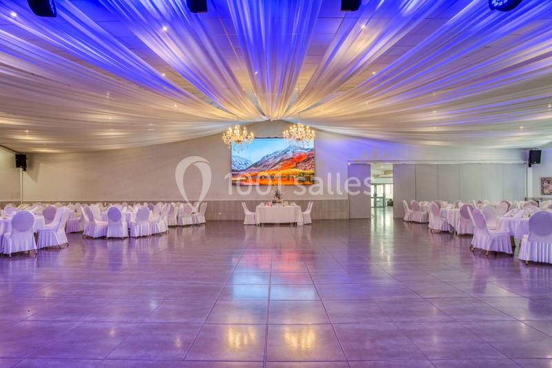 Salle de réception spacieuse avec plafond décoré de drapés lumineux, tables rondes dressées et lustres suspendus.