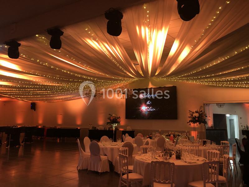 Salle de réception décorée avec des guirlandes lumineuses au plafond, tables dressées et écran affichant des prénoms.