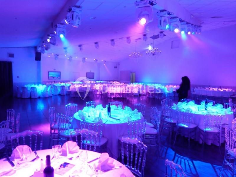 Salle de réception éclairée de lumières violettes, avec tables rondes dressées et chaises transparentes.
