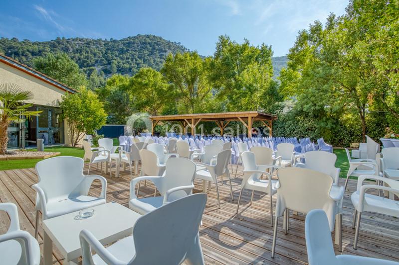 Terrasse extérieure avec chaises blanches, tables et pergola en bois, entourée de verdure et de collines en arrière-plan.