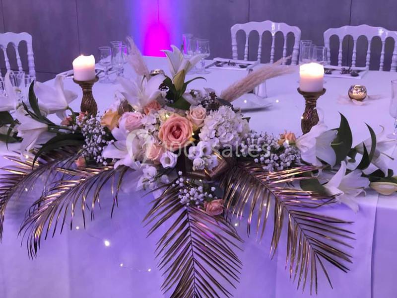 Centre de table floral avec roses, lys, gypsophiles et feuilles dorées, entouré de bougies allumées.