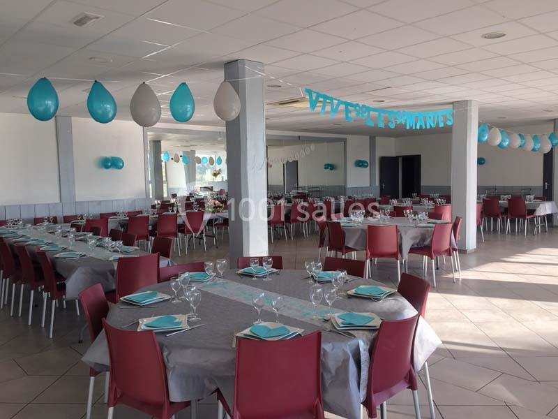 Salle décorée pour un mariage, avec des tables dressées, des chaises rouges et des guirlandes bleues suspendues.