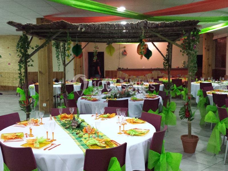 Salle décorée pour un événement, avec des tables rondes ornées de nappes blanches, chaises rouges et décorations tropicales.