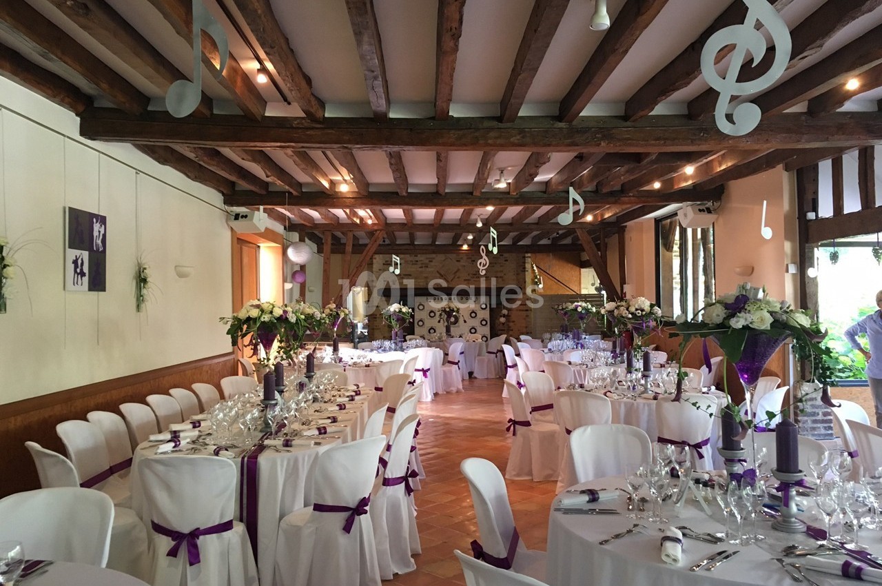Salle de réception décorée avec des tables dressées, des chaises blanches ornées de rubans violets et des notes de musique…