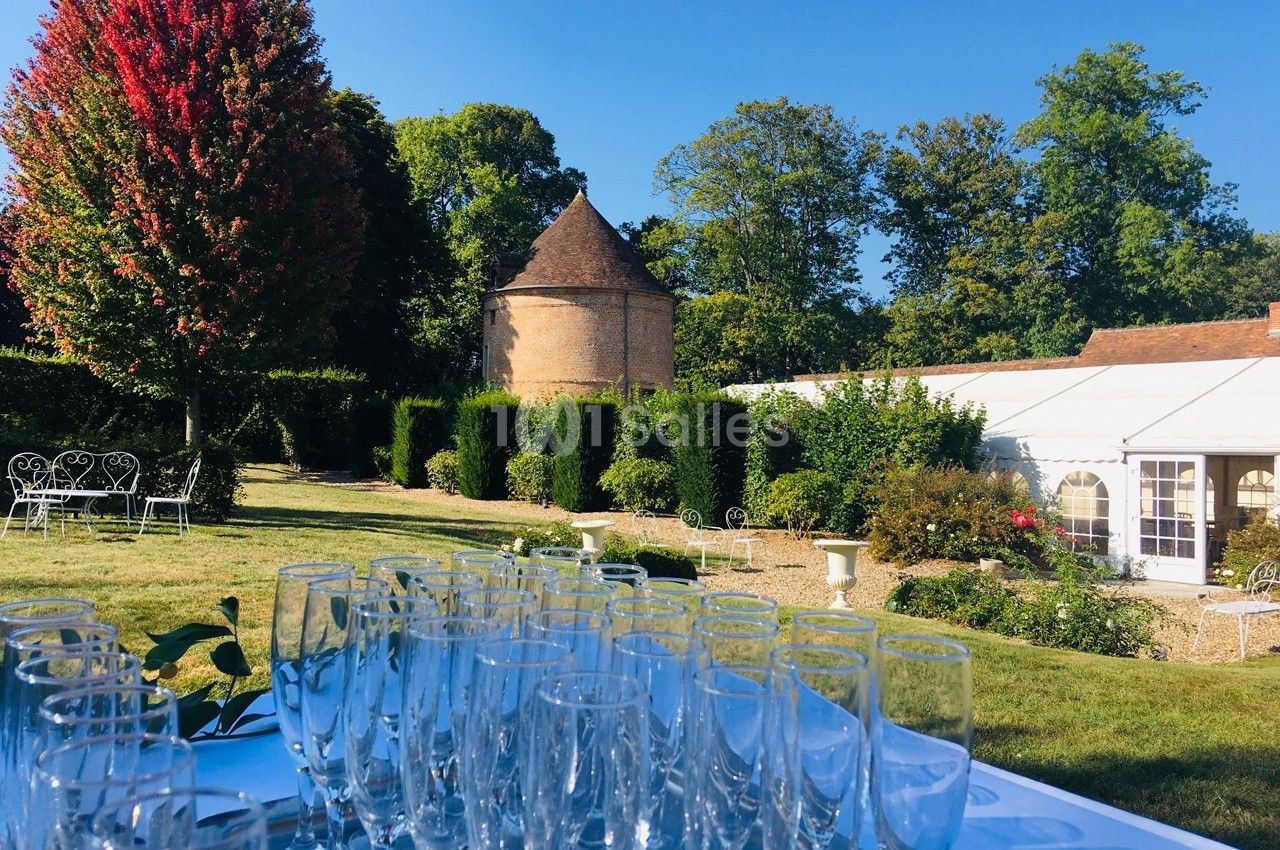 Table avec des verres vides en extérieur, devant un jardin verdoyant, un bâtiment en briques et une tente blanche.