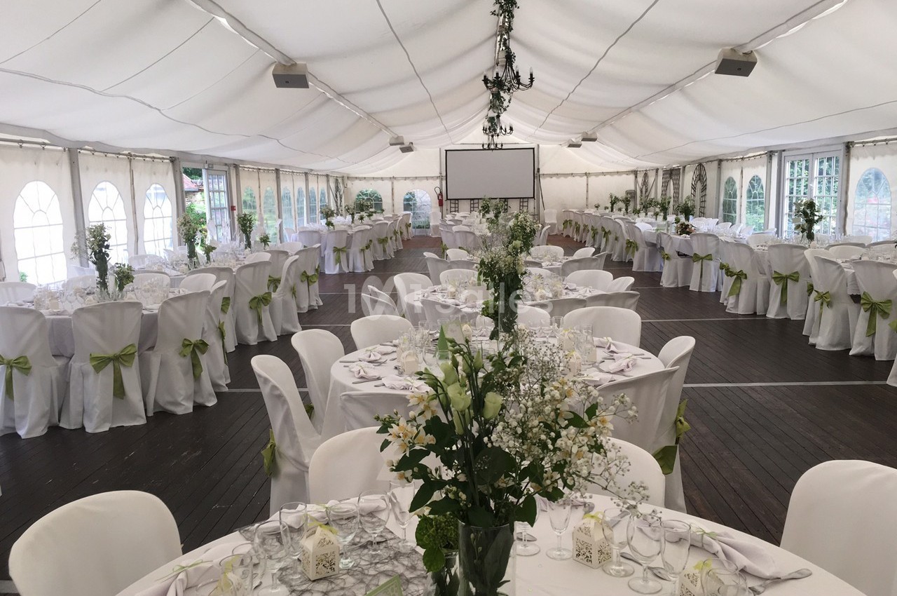 Salle de réception sous chapiteau, tables rondes dressées avec nappes blanches et décorations florales vertes.