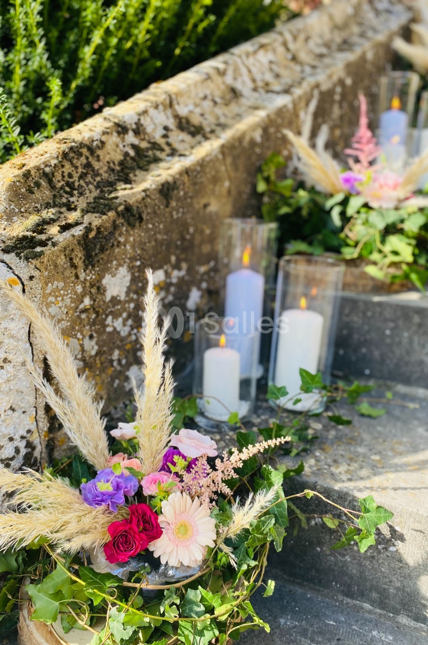 Composition florale avec roses, gerberas et herbes de la pampa, disposée sur des marches en pierre avec des bougies allumées.