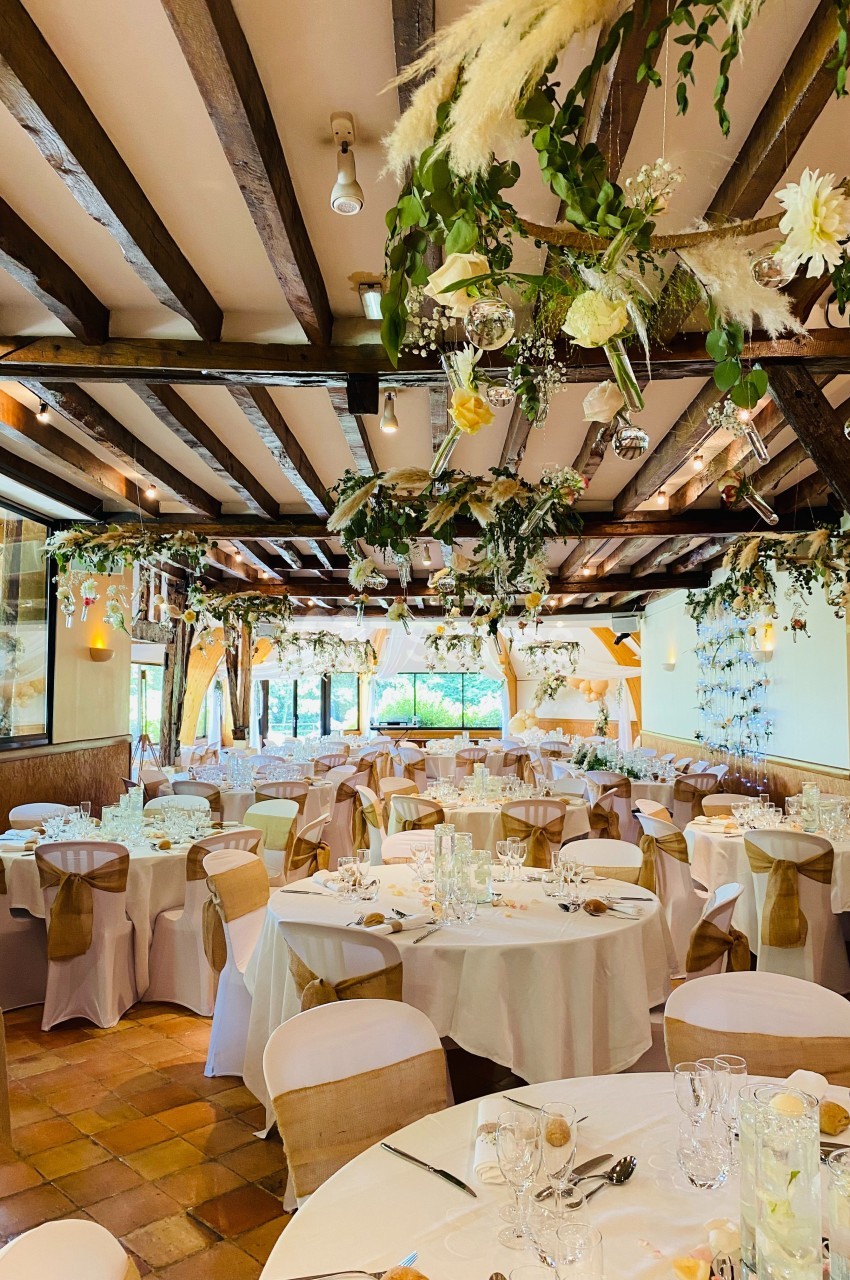 Salle de réception décorée avec des tables rondes, nappes blanches et ornements floraux suspendus.