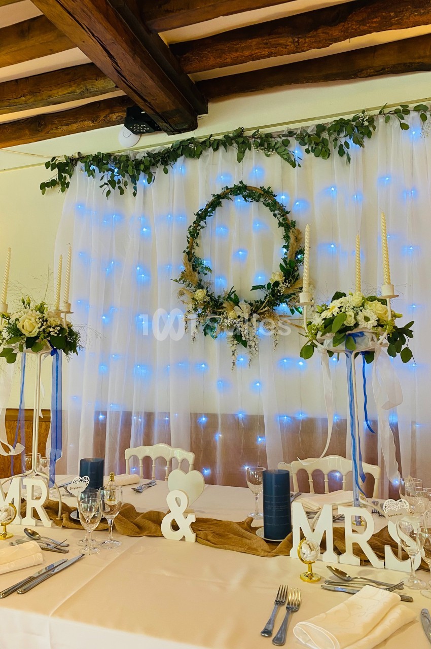 Table de mariage décorée avec des fleurs, bougies, et lettres ’MR & MRS’, devant un fond lumineux et végétal.