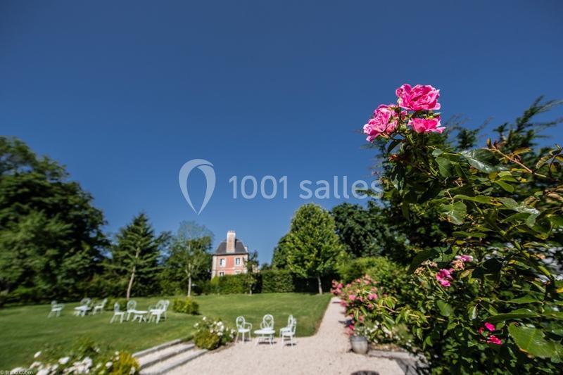 Allée bordée de rosiers roses menant à un jardin avec des chaises blanches et une maison en arrière-plan.