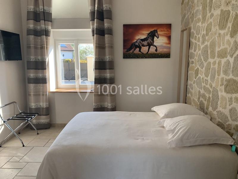Chambre lumineuse avec lit double, mur en pierre, tableau de cheval, fenêtre et télévision fixée au mur.