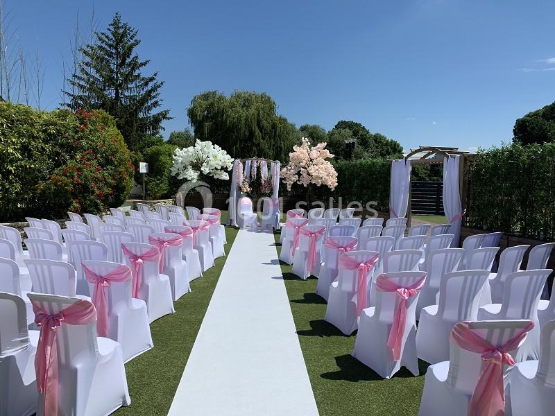 Allée centrale bordée de chaises blanches décorées de rubans roses, menant à une arche fleurie en extérieur.