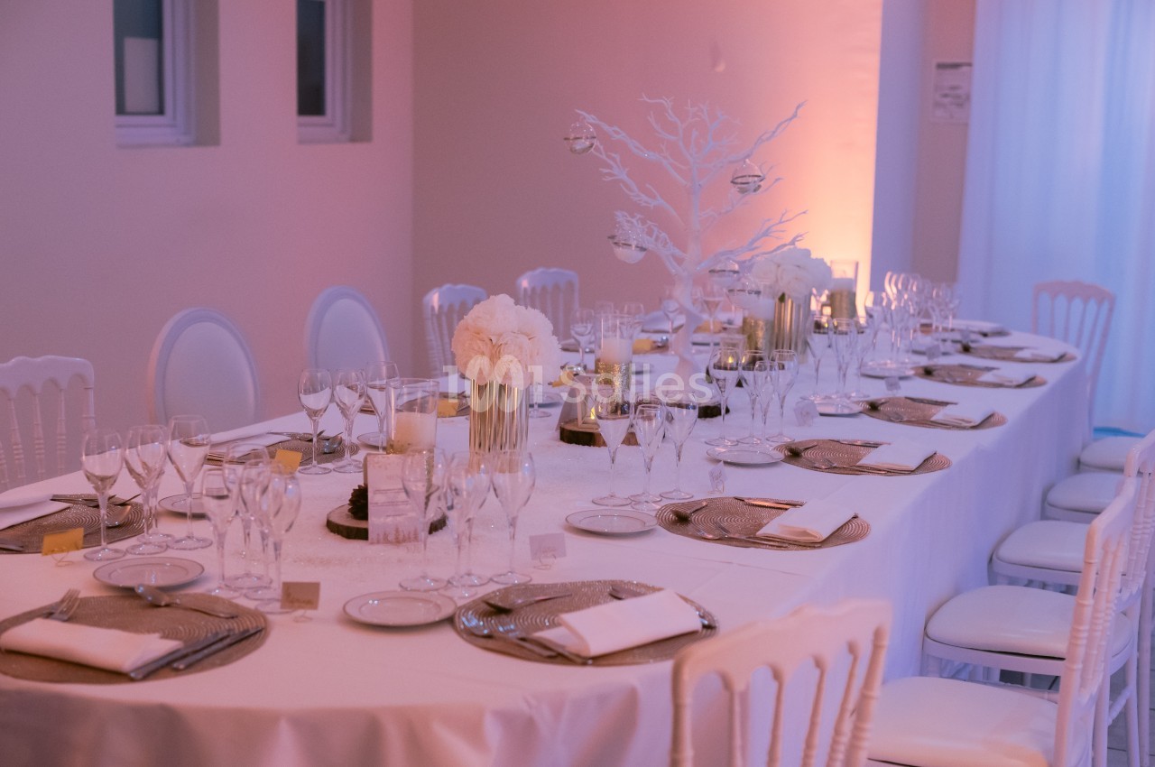 Table élégamment dressée avec nappes blanches, verres, assiettes, serviettes et décorations florales dans une salle éclairée.
