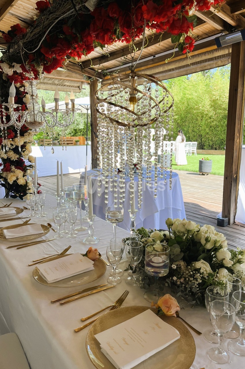 Table élégamment dressée avec vaisselle dorée, chandeliers, fleurs blanches et rouges sous une pergola en bois.