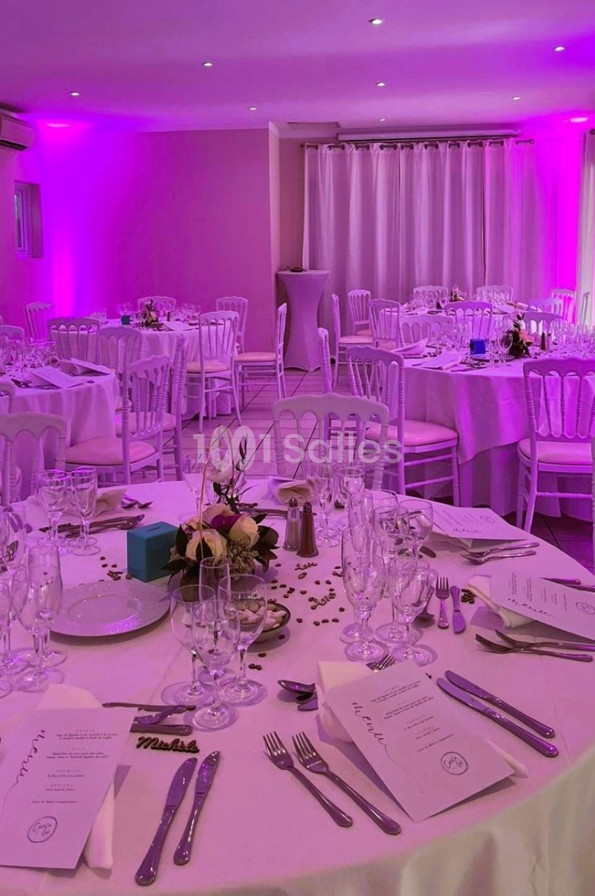 Salle de réception décorée avec des tables rondes dressées, éclairage rose et chaises blanches.