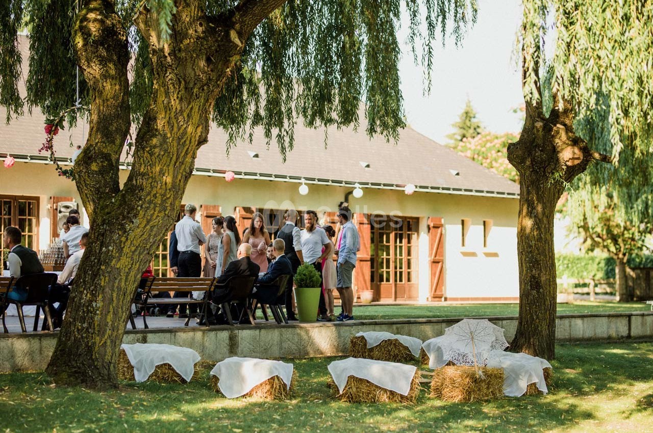 Groupe de personnes discutant près d'un bâtiment, avec des bottes de paille décorées au premier plan sous des arbres.