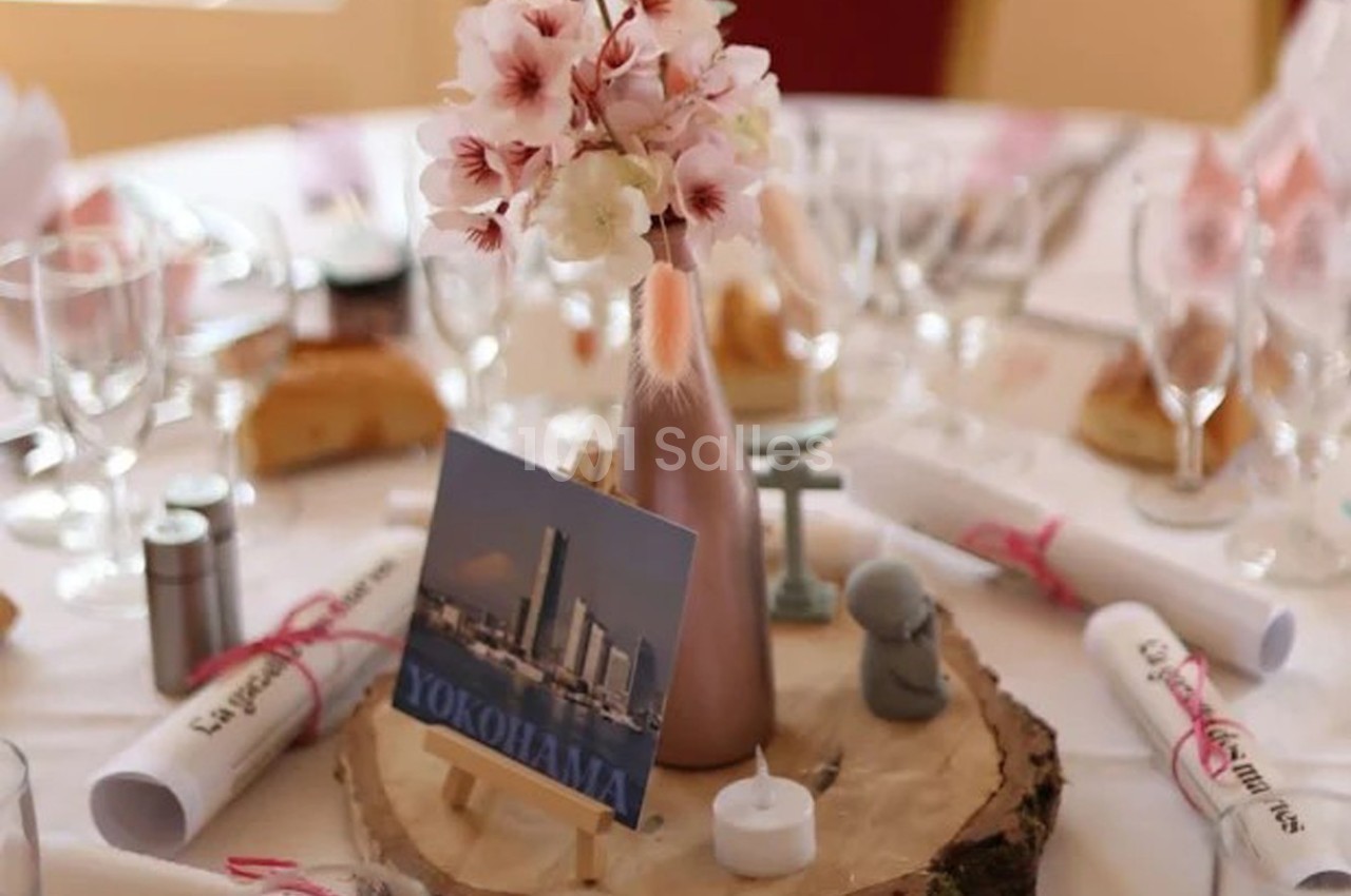 Centre de table avec vase rose, fleurs, carte ’Yokohama’, bougie et rondin de bois, entouré de verres et serviettes.