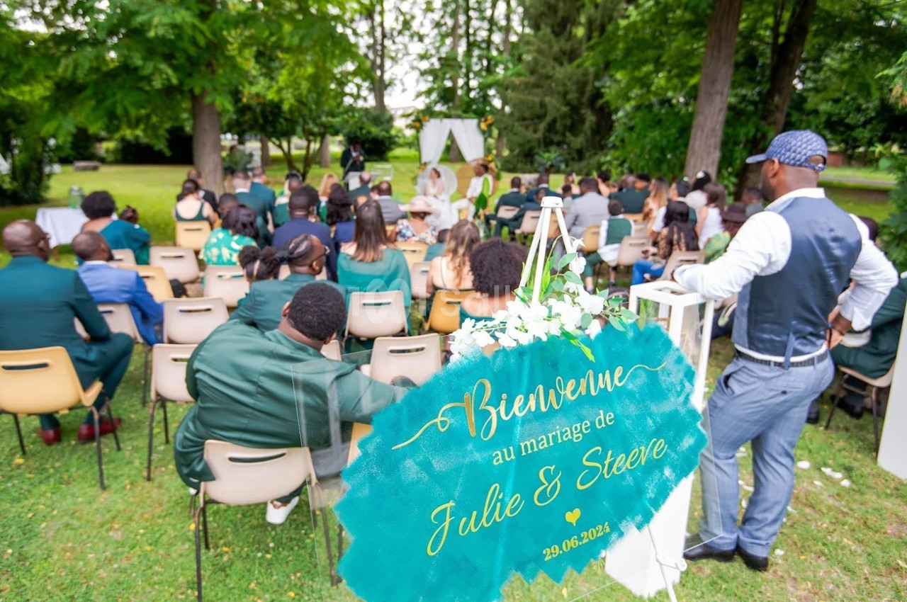 Invités assis en plein air lors d'une cérémonie de mariage, avec un panneau de bienvenue au premier plan.