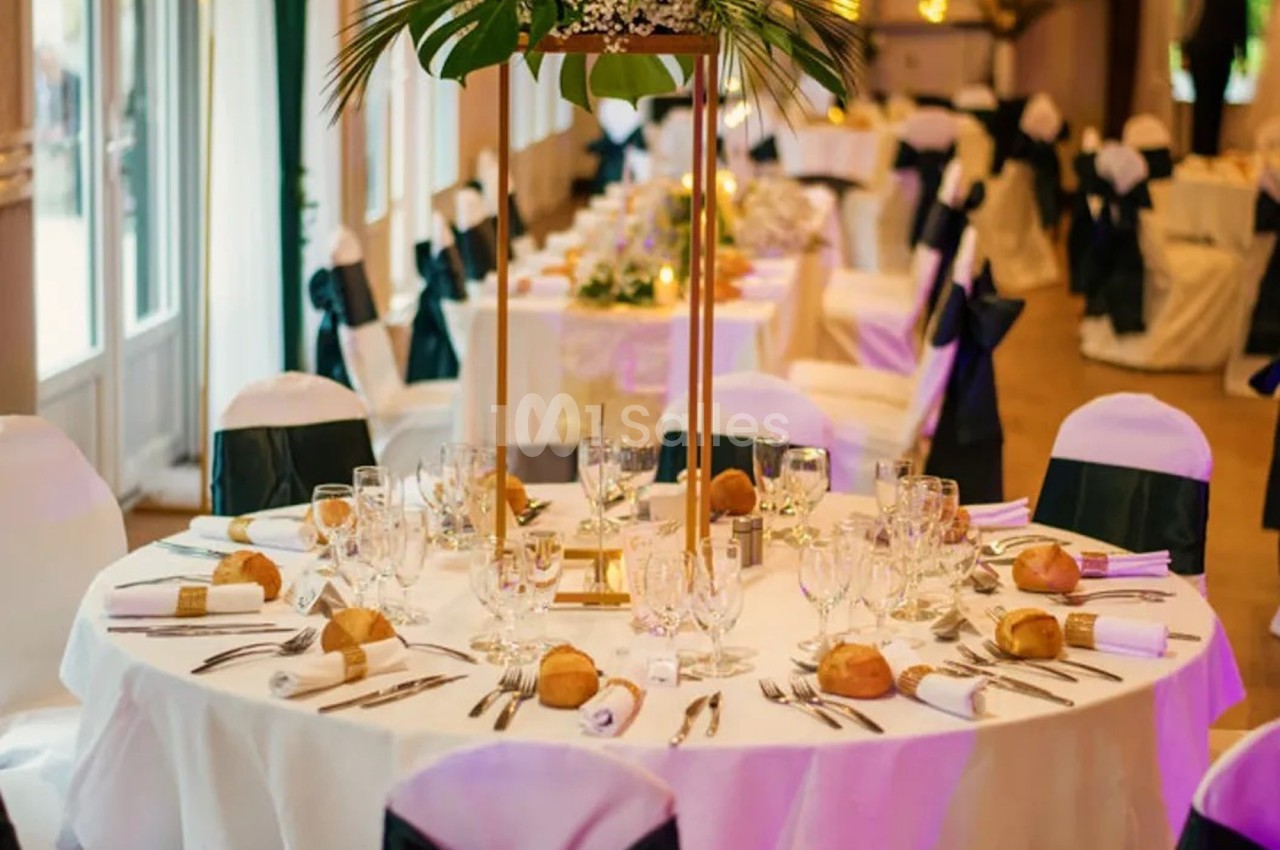 Salle de réception décorée avec des tables rondes dressées, nappes blanches, couverts élégants et centres de table floraux.