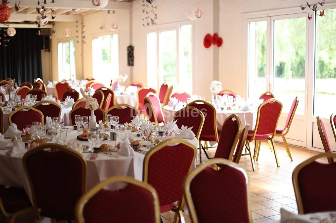 Salle de réception lumineuse avec des tables rondes dressées, nappes blanches et chaises rouges.