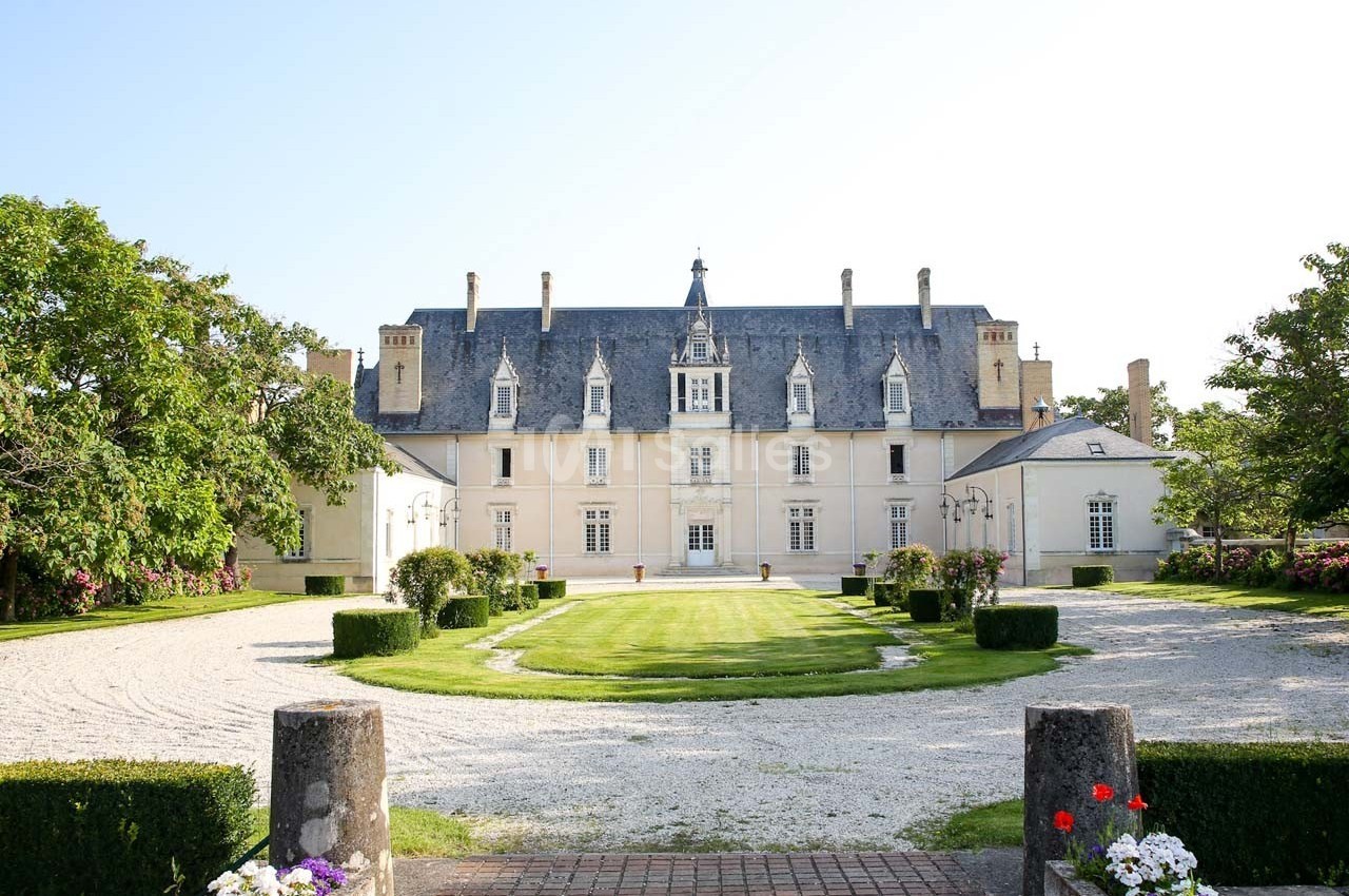 Façade d'un château blanc entouré de jardins, avec une allée gravillonnée et des arbres en arrière-plan.